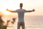 Person mit ausgebreiteten Armen, der dem Meer bei Sonnenuntergang zugewandt ist