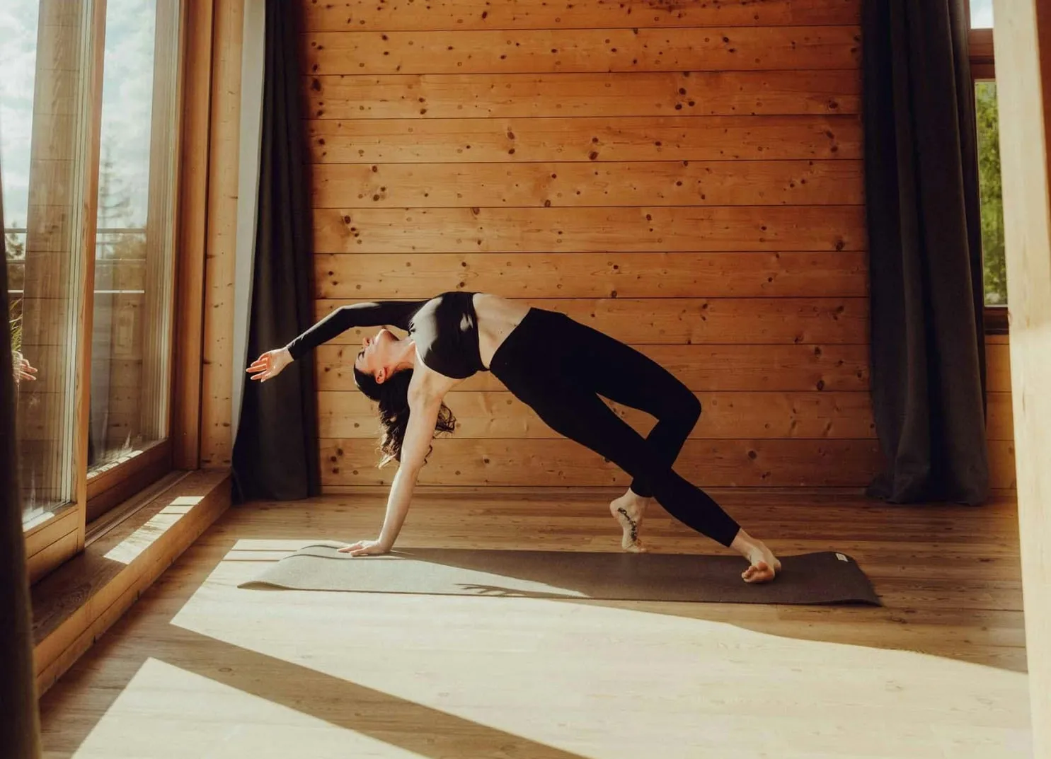 Eine Frau in einem Holzinterieur mit großen Fenstern, die eine Yoga-Pose ausführt